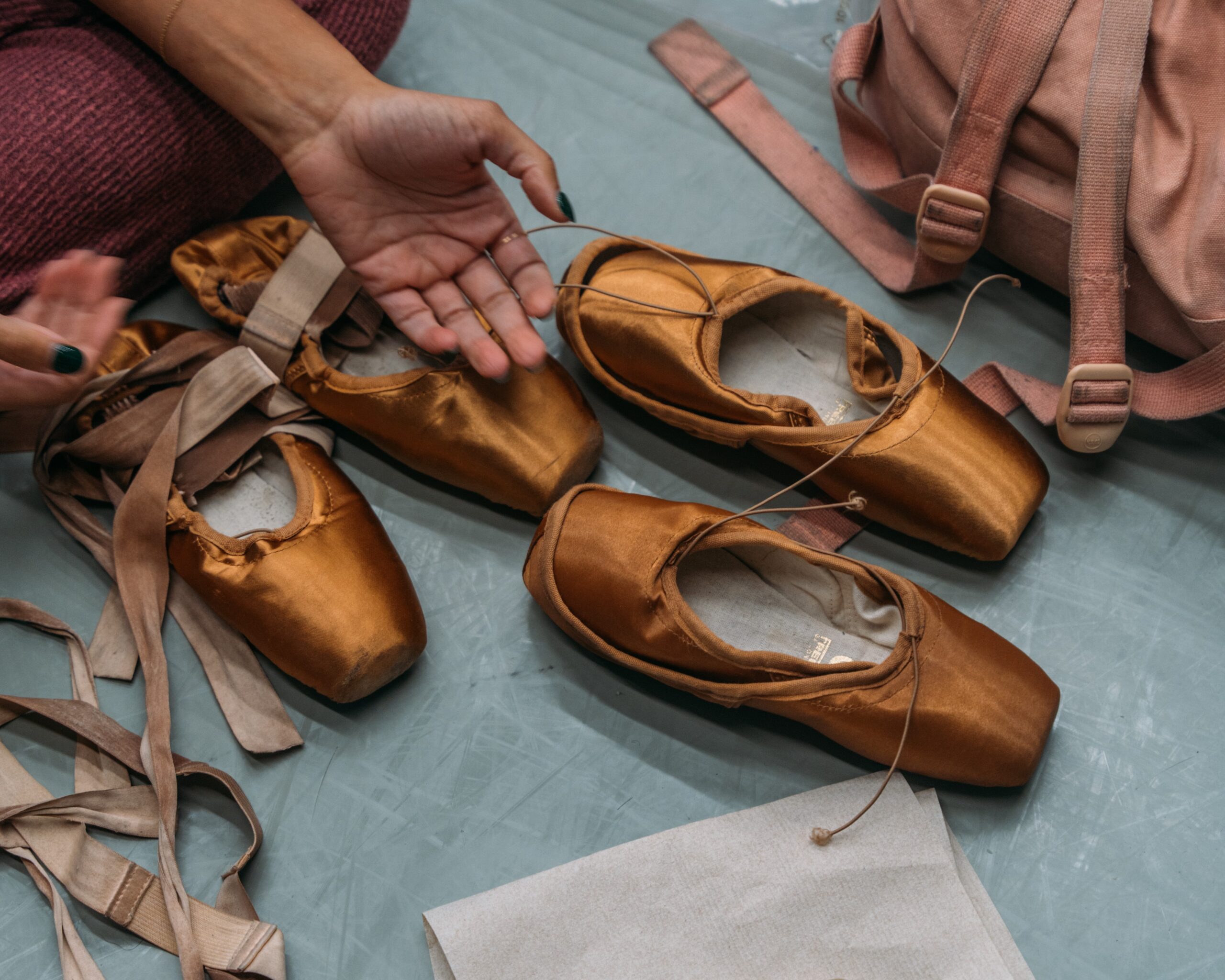 Multiple pairs of pointe shoes on the floor, along with paper towels, and a pink backpack