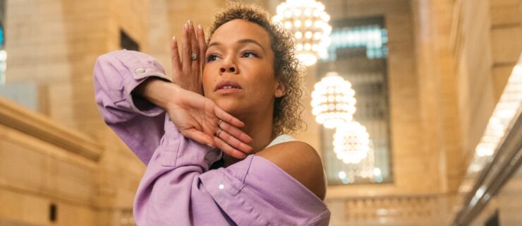 a female dancer wearing a purple button down shirt and jeans dancing in a large open room. she poses with her arms crossed and her hands near her face