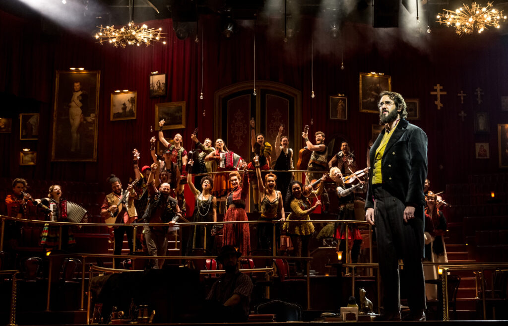 a male performer downstage looking sullen with a large group of performers standing behind him cheering 