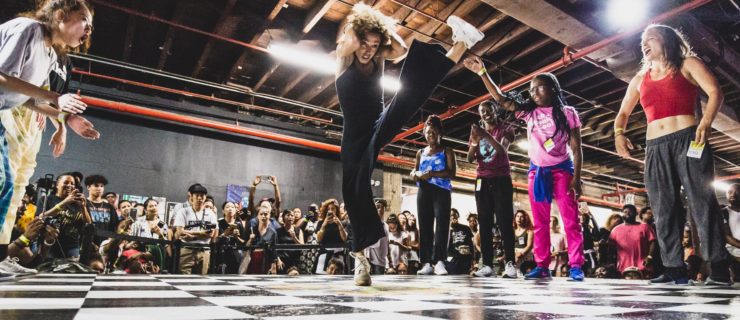 A dancer jumps and kicks one leg to her head in a battle on a grid-like floor