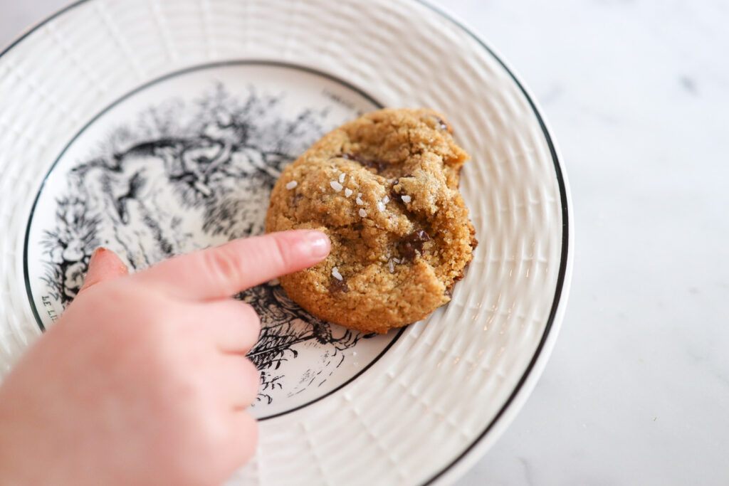 A child's finger points at a chocolate chip cookie on a black and white plate