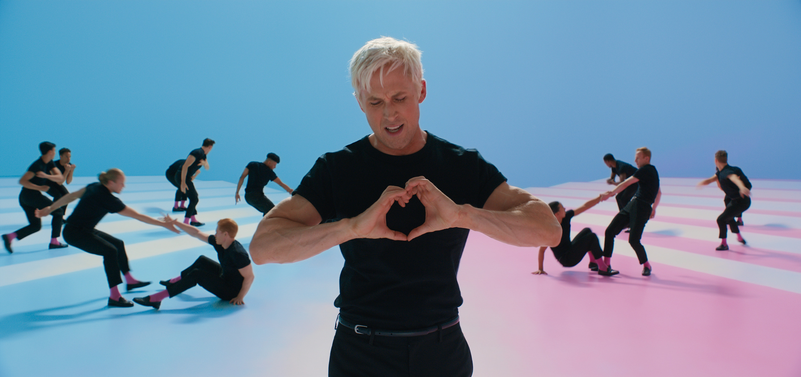 Gosling, wearing a black tee and jeans, makes a heart with his hands in front of his chest. An ensemble of identically-dressed men dance behind him.