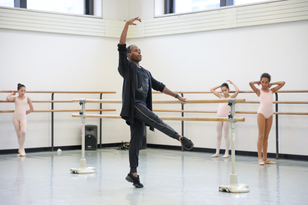 McDaniel, a dark-skinned man wearing black rehearsal clothes and dance sneakers, leads a classroom of young students in pink leotards at SAB.