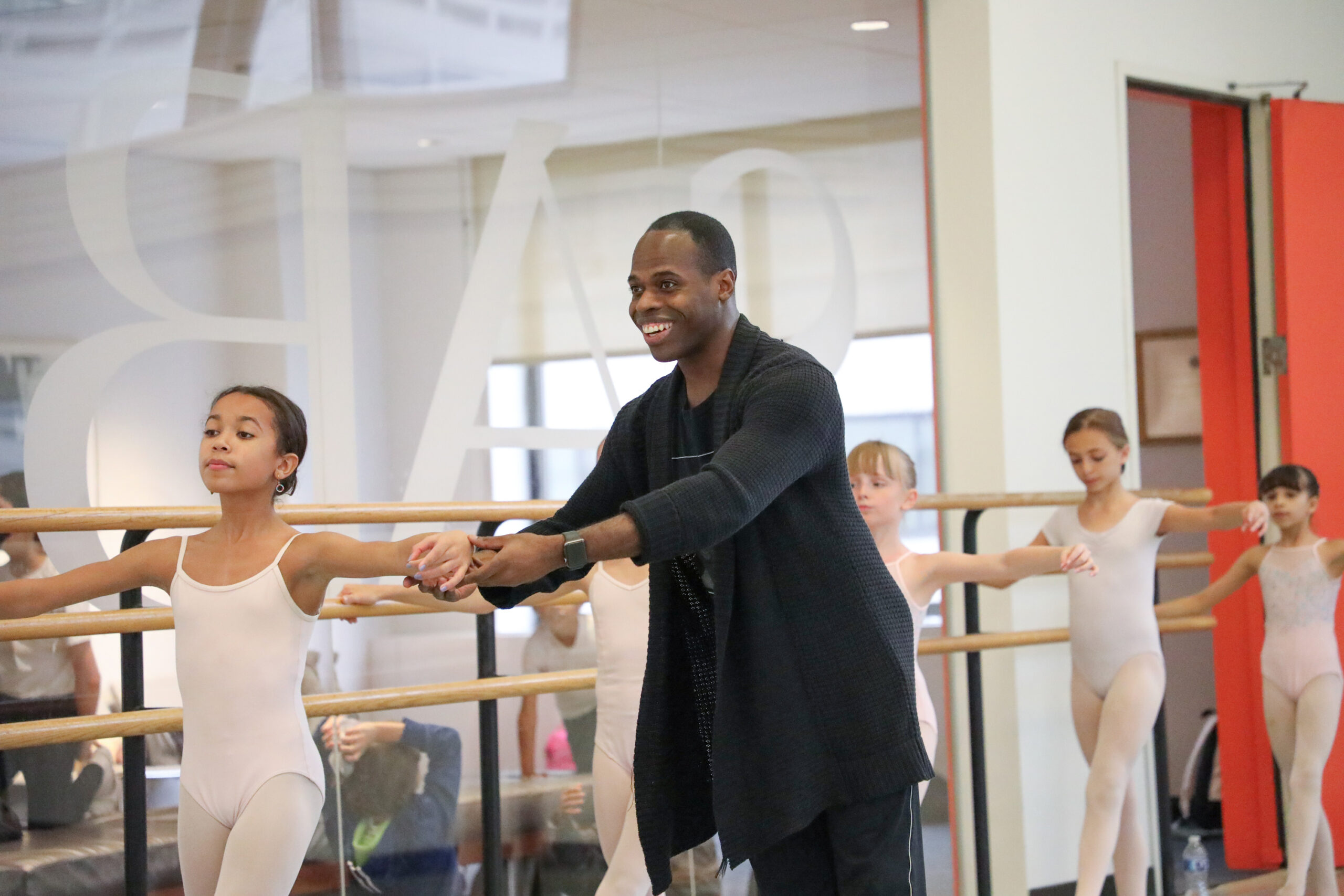 McDaniel, a dark-skinned man wearing black practice clothes, smiles as he corrects the arm placement of a young student in a ballet classroom.