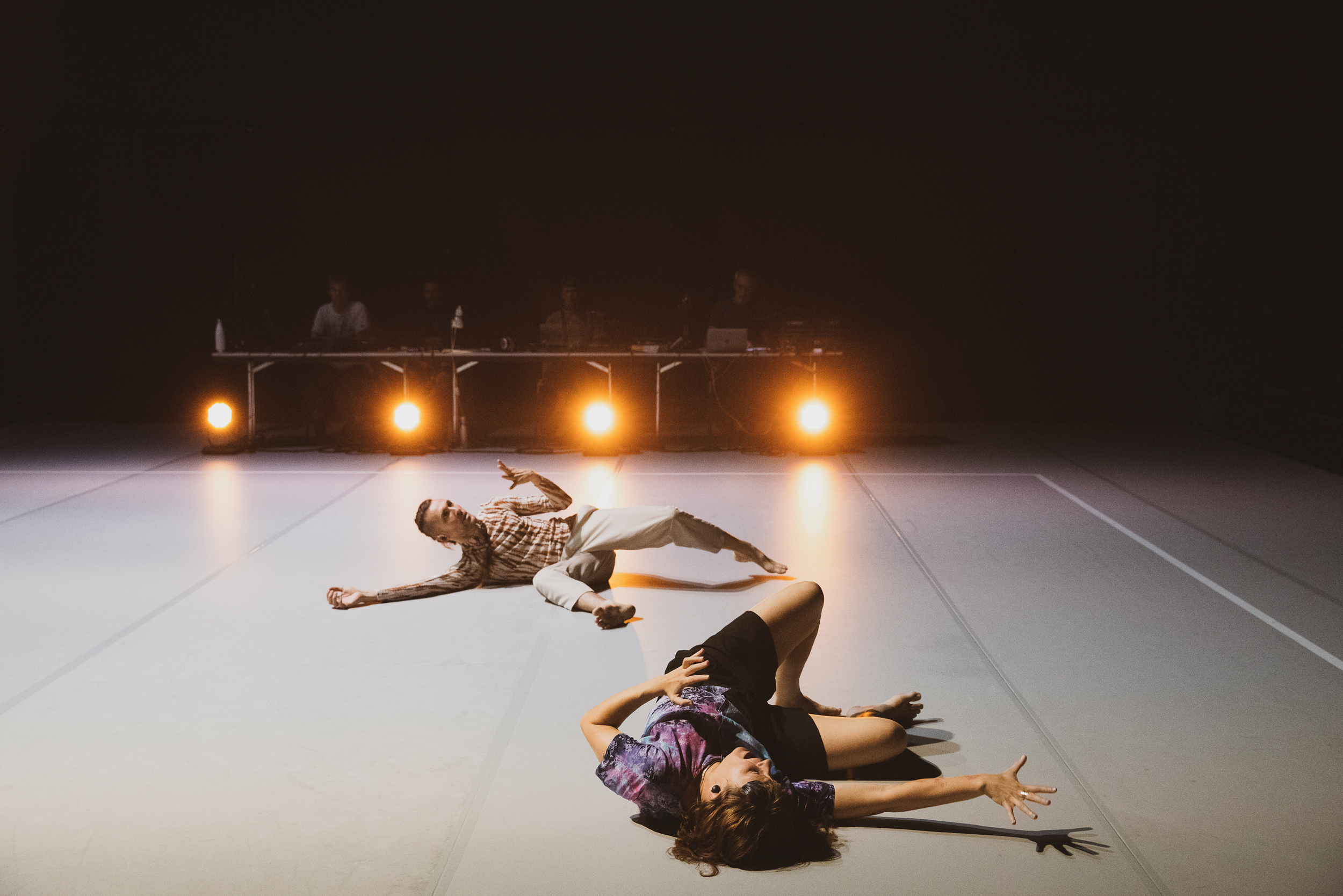 Obergfell and Delorme writhe on a gray dance floor, illuminated by four bright floor lights. In the blurred background, four shadowy figures sit at a table strewn with wires and laptops.