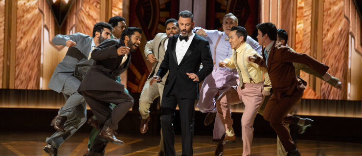 On the gilded Oscars stage, a group of eight male dancers in suits surround Jimmy Kimmel, who wears a black tuxedo, as they energetically perform the “Naatu Naatu” hook-step dance move. Kimmel smiles and looks down as he dances along.