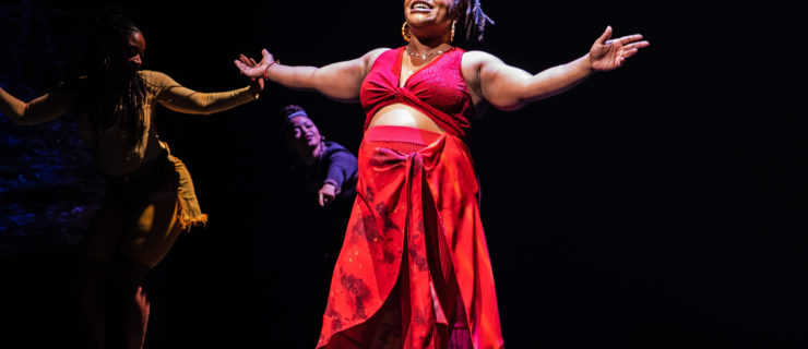 Kenita R. Miller, a Black pregnant Broadway star, extends her arms wide and beams onstage. She is wearing a red crop top and pants, and her pregnant belly is visible.