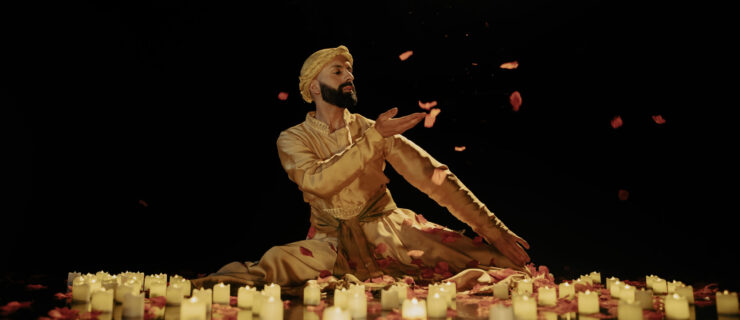 Aakash Odedra sits with his legs folded beneath him amidst lit candles and falling flower petals. One hand crosses his chest, palm upraised as though to catch a falling petal. He looks over his shoulder, gaze downturned. He wears a gold head covering that matches his costume.