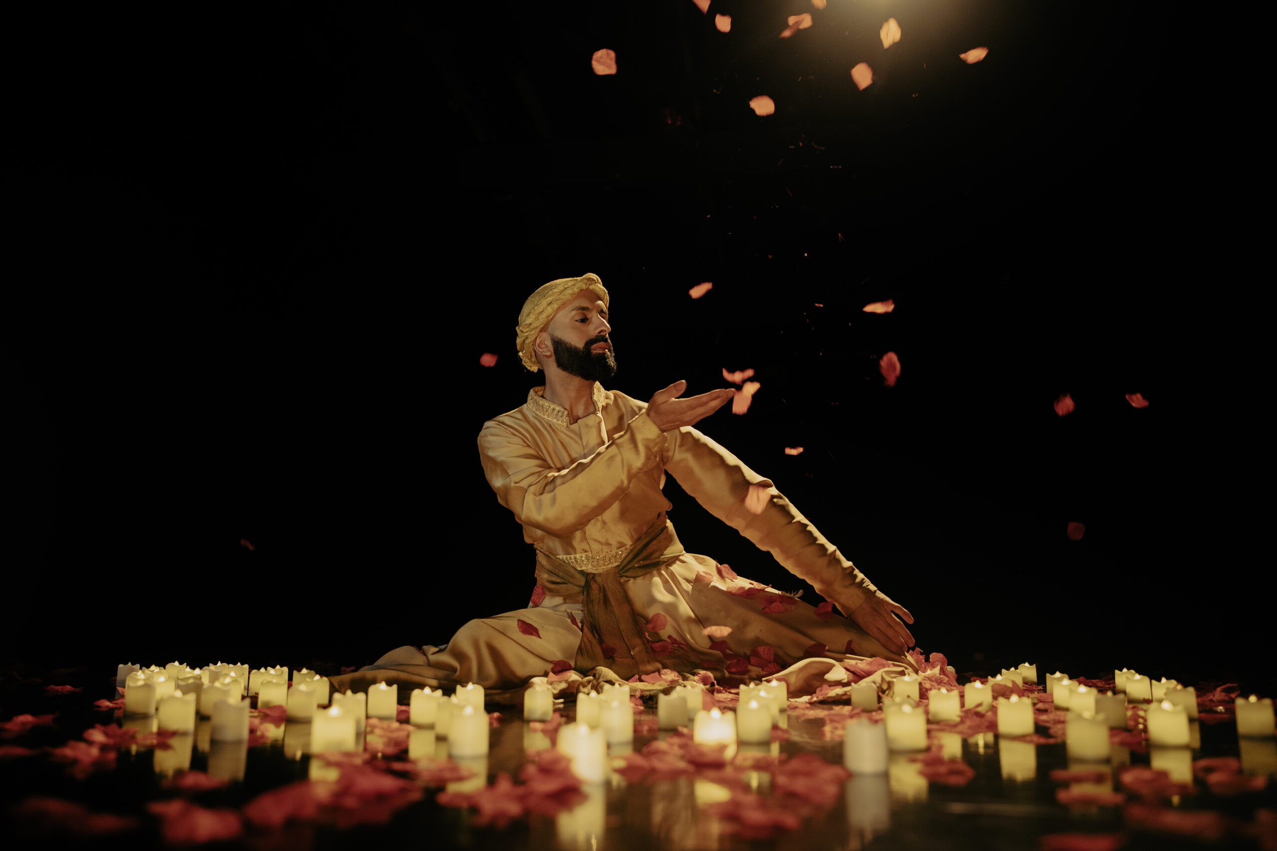 Aakash Odedra sits with his legs folded beneath him amidst lit candles and falling flower petals. One hand crosses his chest, palm upraised as though to catch a falling petal. He looks over his shoulder, gaze downturned. He wears a gold head covering that matches his costume.