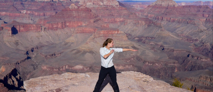 Denton, wearing a loose white shirt and black pants, stands at the rim of the Canyon on a brilliantly sunny day, her legs planted in a wide second position, looking out over her extended left arm.