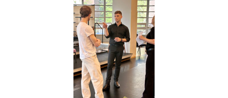 Three people stand in a sunlit dance studio. Danchig-Waring, in white practice clothes, watches attentively as Novak, in all black, gestures toward him with his right hand; McCann, also in all black, looks on.