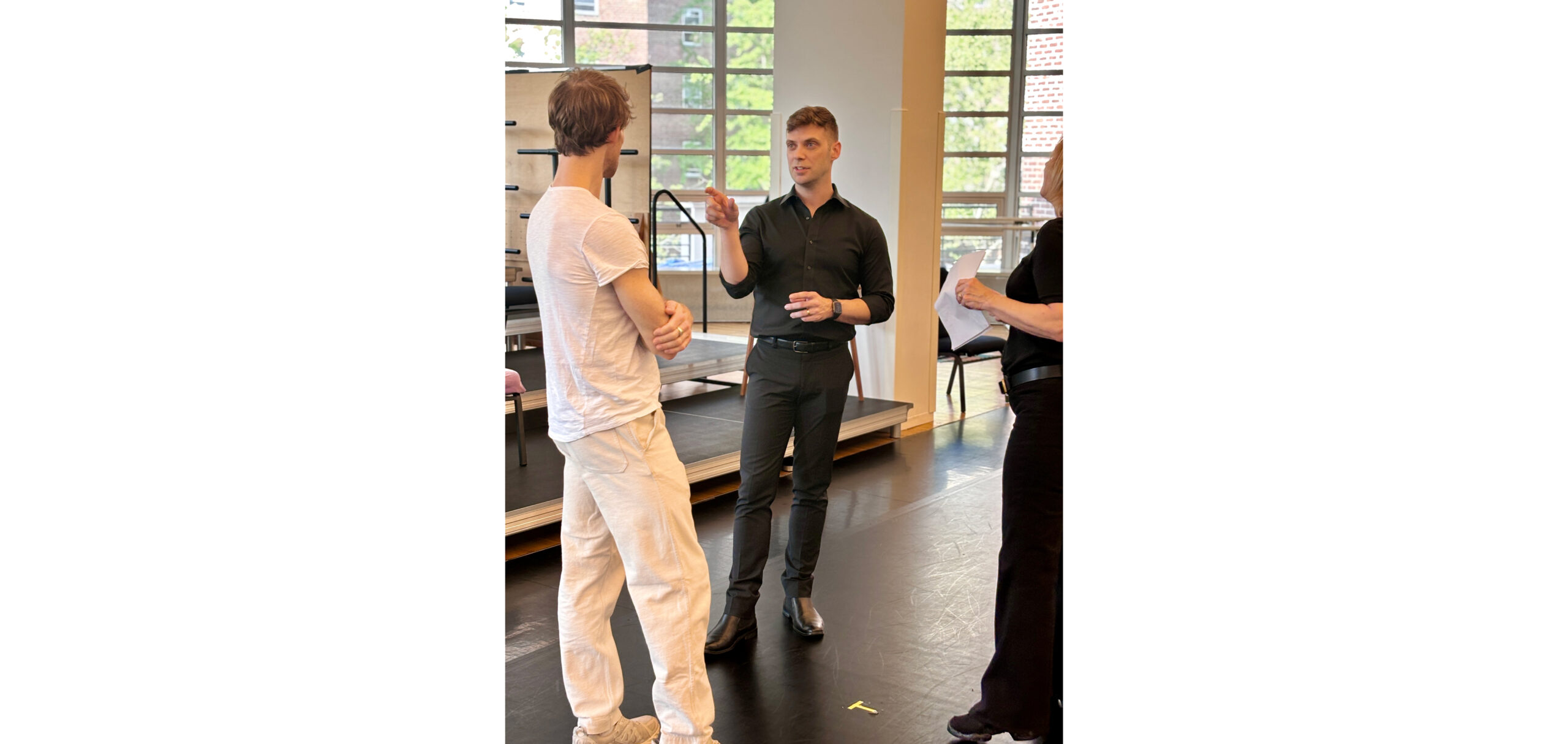 Three people stand in a sunlit dance studio. Danchig-Waring, in white practice clothes, watches attentively as Novak, in all black, gestures toward him with his right hand; McCann, also in all black, looks on.