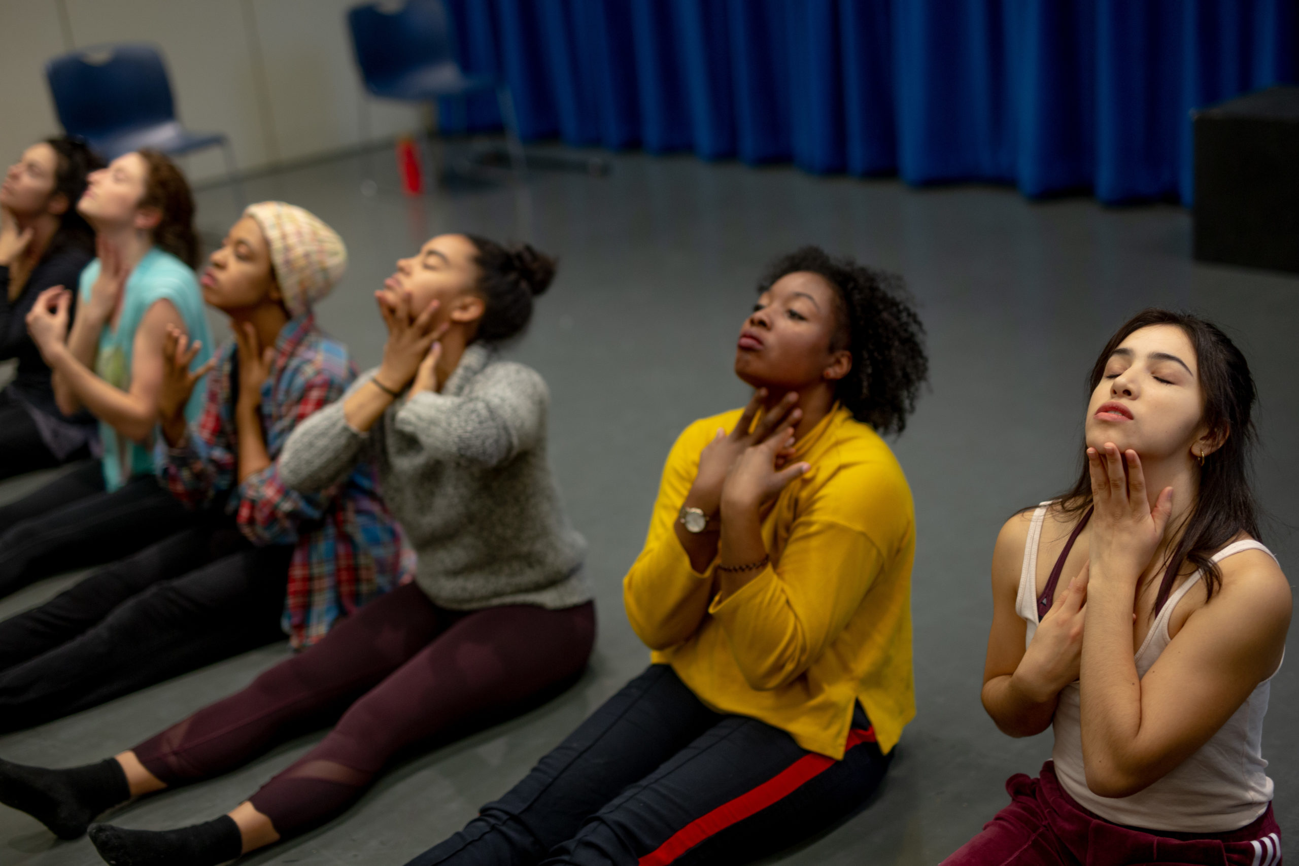 A line of women sit in a horizontal line with their legs stretched out in front of them. THeir eyes close as they bring both hands to their throats, heads slightly tipped back.