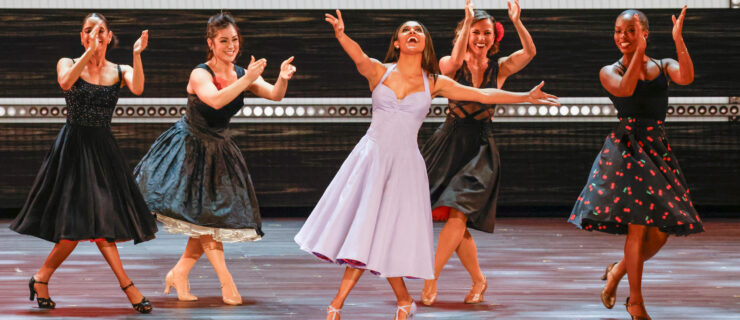 DeBose, in a swirly lilac dress, poses onstage in front of four other clapping dancers in similarly full-skirted black dresses.