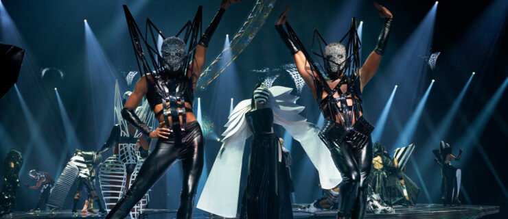 A stageful of dancers pose in elaborate, futuristic black-and-white costumes with painted or bejeweled full-face masks.