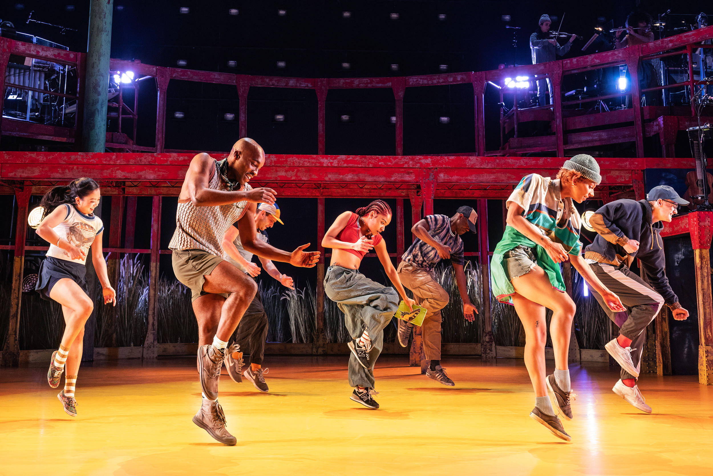 A collection of dancers in colorful street clothes and sneakers are captured onstage mid-jump, with their right knees slightly bent and their arms floating loosely. In the background, red scaffolding looms above the golden floor.