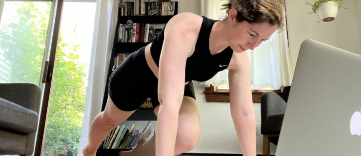 Heimlich, wearing a black sports bra and bike shorts, is seen in her living room with her laptop on the floor in front of her. She looks at the screen as she executes a modified plank position, with her left leg raised in a parallel passé.