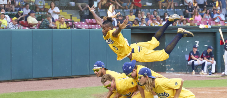 Five men in matching bright yellow baseball uniforms pose together on the field. Four of them crouch with their hands on their knees, while a fifth, Maceo Harrison, flies parallel to the ground over their backs, knees bent behind him and arms splayed out at his sides.