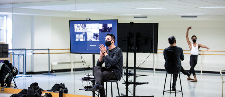 An Asian man sits on a stool at the front of a ballet studio, wearing a mask, clapping his hands together.