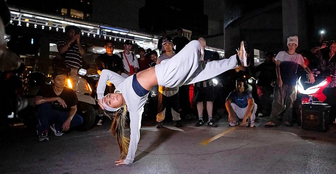 Logan "Logistx" Edra in a cypher. She is captured balancing on one hand, face up, body parallel to the floor. Her other hand touches her head; one leg bends over top the other as it kicks out straight.