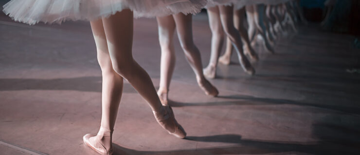 A line of dancers in white tutu are shown from the waist down, each one in pointe shoes and tights with one foot tucked behind the other