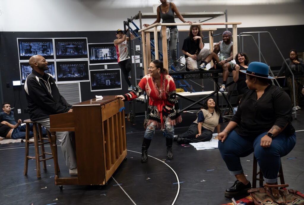 a group of performers surrounding a man at a piano in rehearsal 