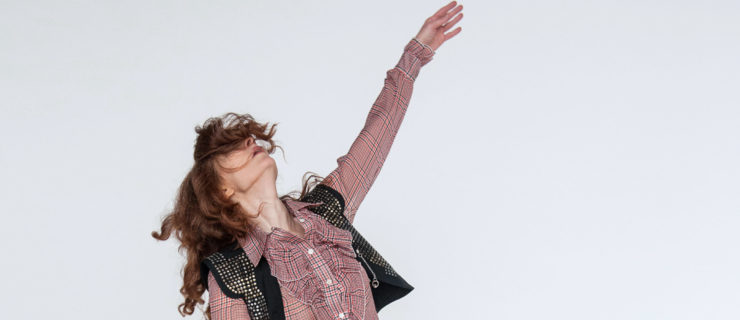 woman in yellow pants and plaid shirt, one arm up one foot in coupe