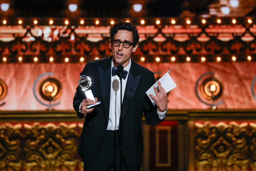 Peck, wearing a tuxedo, speaks at a microphone onstage, holding his Tony Award.