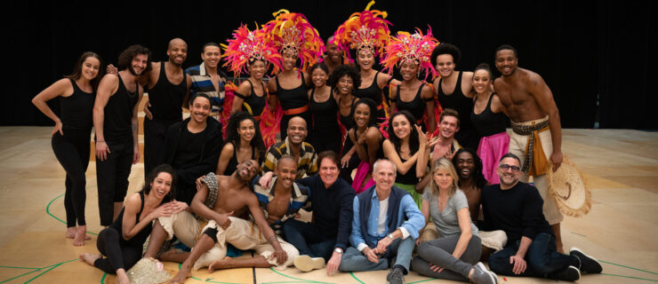 A large group of artists, some wearing flamboyant feathered headpieces, poses on a basketball court.