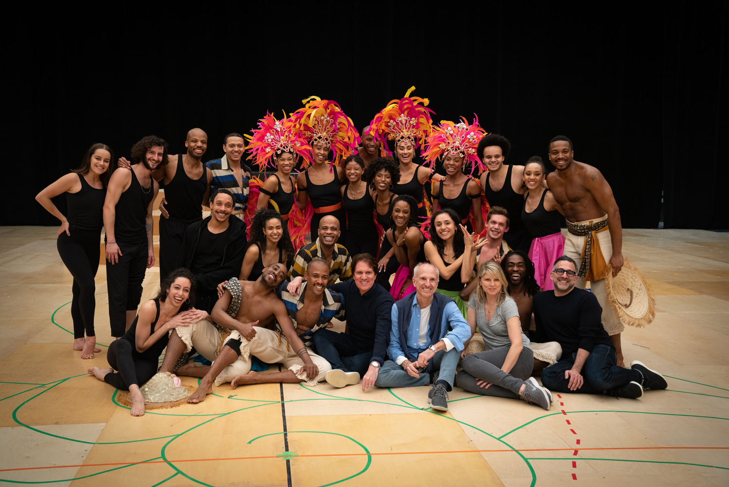 A large group of artists, some wearing flamboyant feathered headpieces, poses on a basketball court.