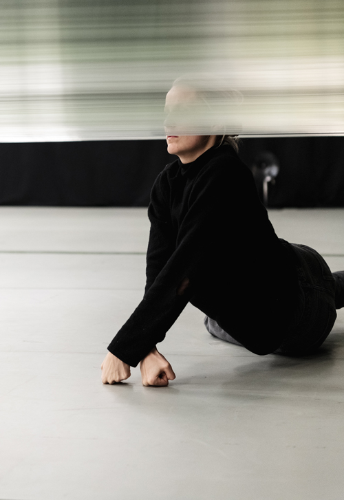 Dahl, in a black turtleneck, lies on the floor, supporting herself with her arms, which are crossed at the wrists and end in balled fists. Her head pokes up through a canopy of fine silver threads.