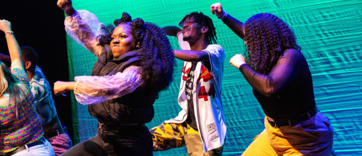 Four dancers in brightly colored costumes are shown from the knees up. In unison, they hike up on knee, the same hand raised in a fist overhead.