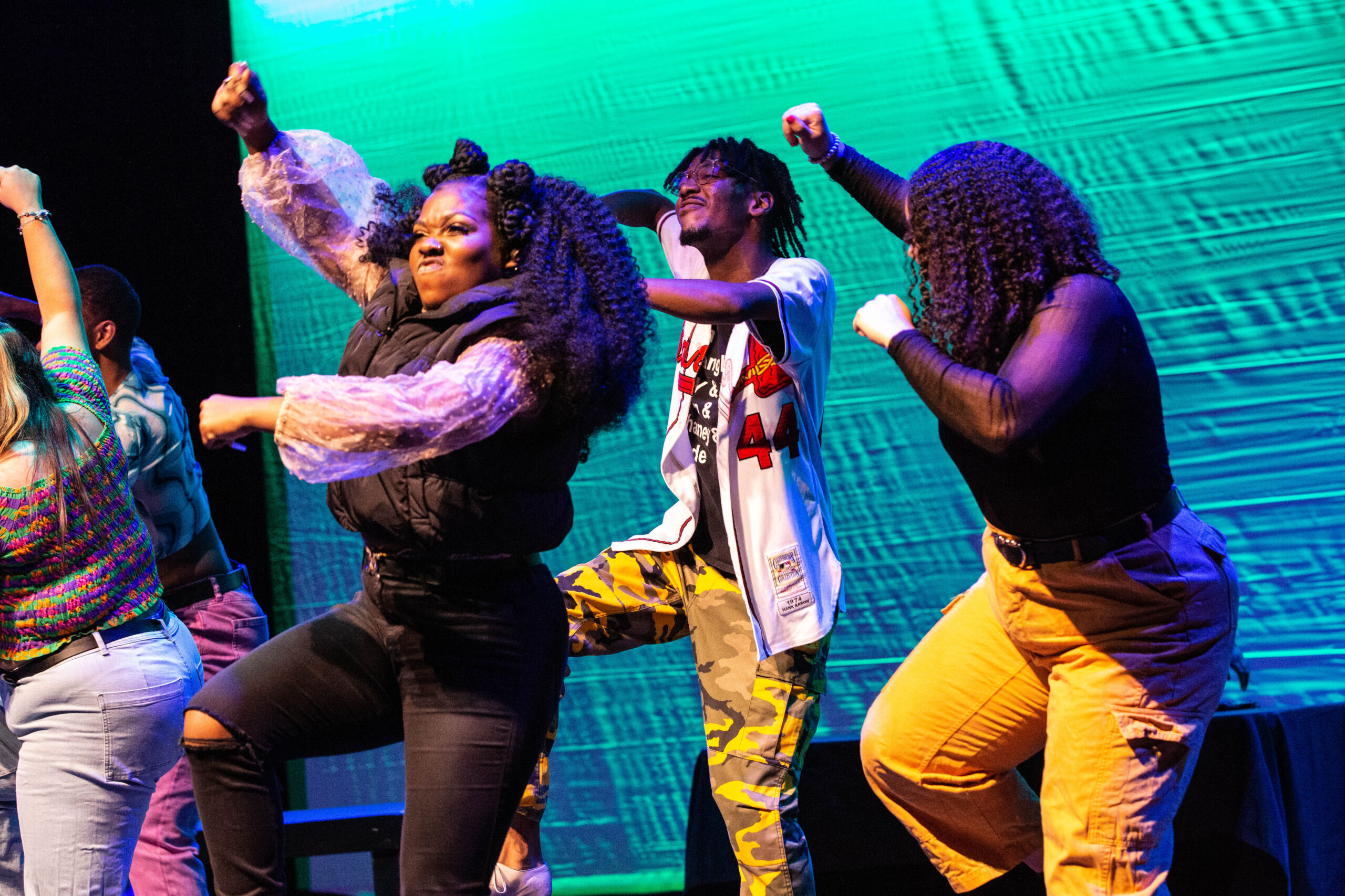 Four dancers in brightly colored costumes are shown from the knees up. In unison, they hike up on knee, the same hand raised in a fist overhead.