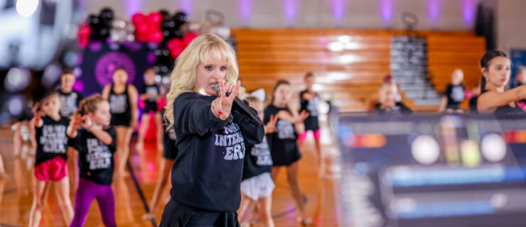 a blonde female instructor talking into a microphone while holding two fingers out in front of her while leading a group of young students