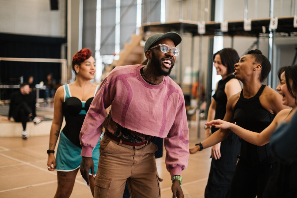 Kelly, wearing a pink cropped sweatshirt and olive baseball cap, laughs as he works with a studio full of dancers.