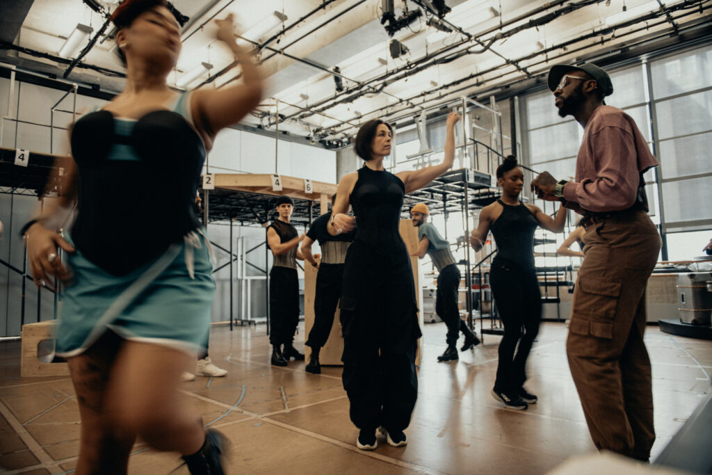 Kelly, wearing a pink cropped sweatshirt and olive baseball cap, watches a studio full of dancers.