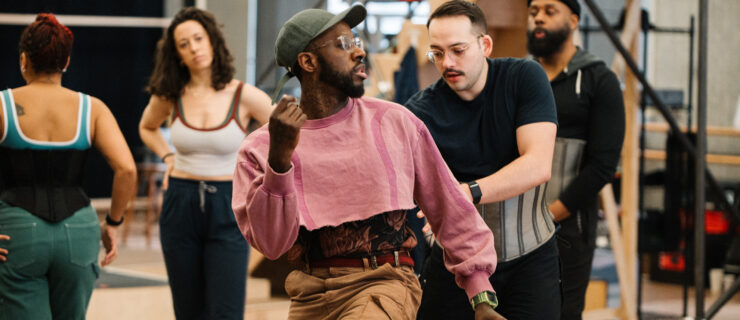 Kelly, wearing a pink cropped sweatshirt and olive baseball cap, demonstrates a step at the front of a studio full of dancers.