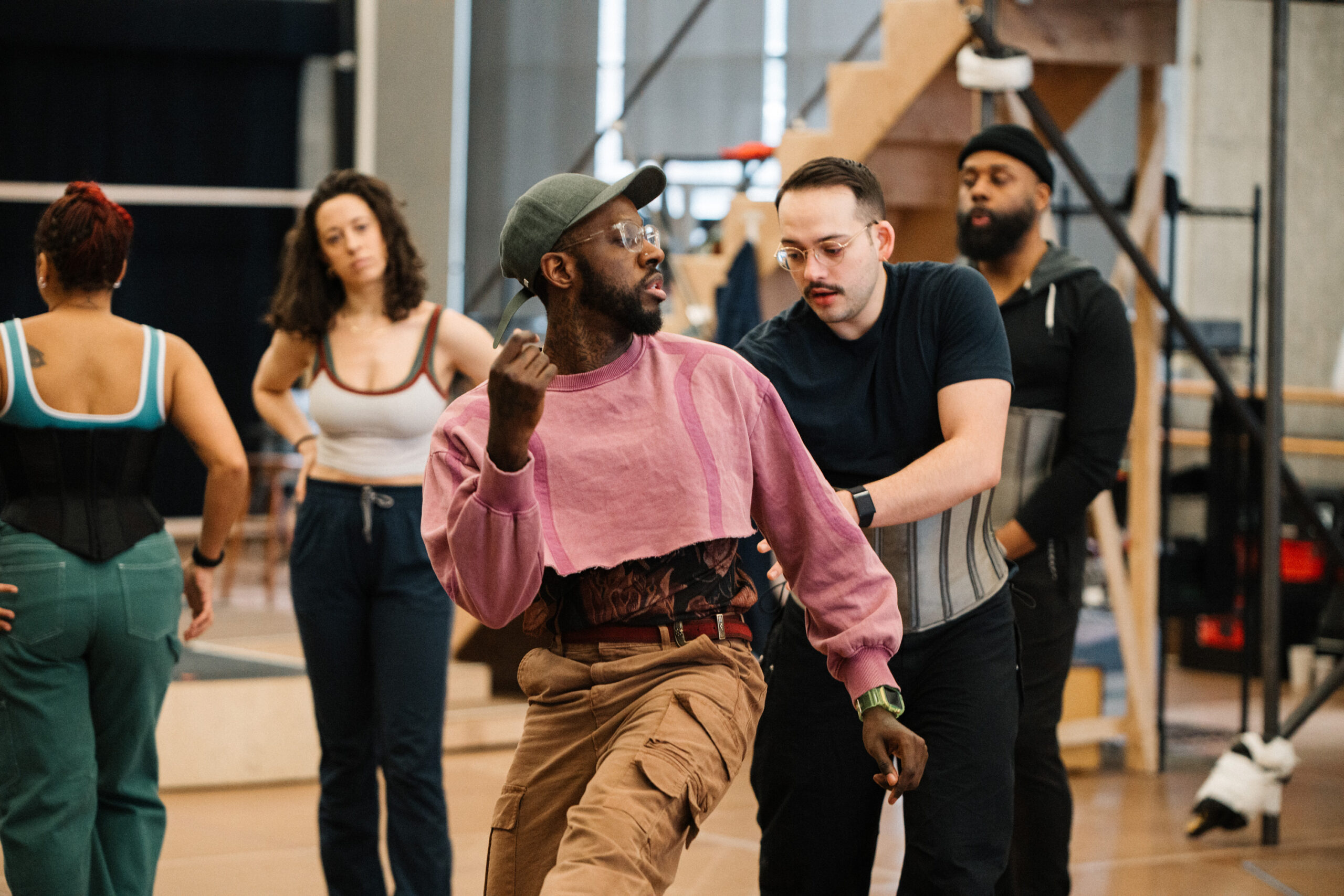 Kelly, wearing a pink cropped sweatshirt and olive baseball cap, demonstrates a step at the front of a studio full of dancers.