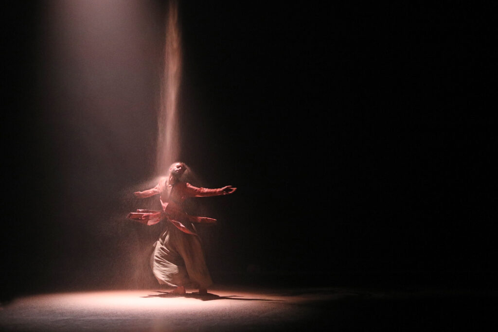 Aakash Odedra turns beneath a spotlight as sand falls from overhead to stream over his shoulders.