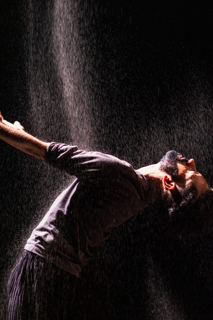 Aakash Odedra bends back in a spotlight, allowing falling sand to stream onto his chest as his arms rise before him.