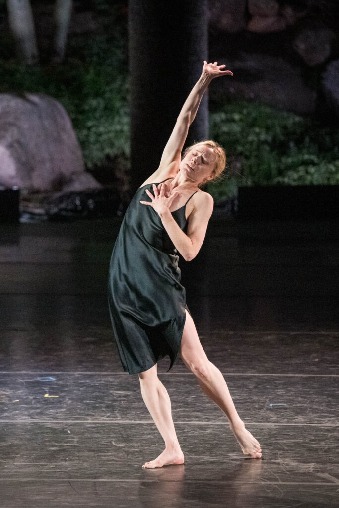 Sara Mearns is barefoot as she leans into one hip and arches away from it, dragging the other behind her. She presses one palm to her chest as the other arm reaches for the sky. She wears a dark green nightgown.