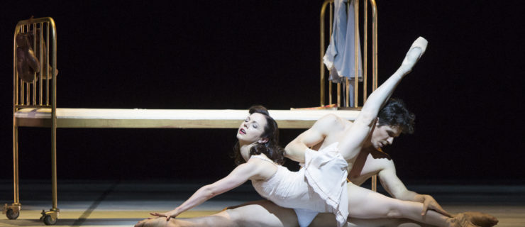 Jillian Vanstone and Guillaume Coté are white ballet dancers with dark hair. They wear delicate white costumes and lie on the stage floor in front of a small bed, facing opposite directions. Vanstone's back is arched and her left leg points back and upward.