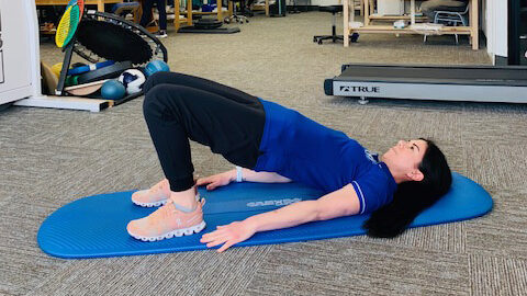 a female physical therapist demonstrating by lifting her hips off the mat in a bridge position 