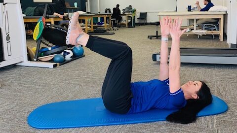 a female physical therapist demonstrating by lying on her back with both arms straight up and legs bent at 90 degrees 