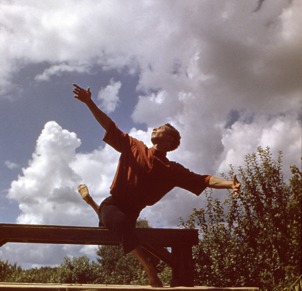 a dancer performing on a bench outside surrounded by trees and clouds