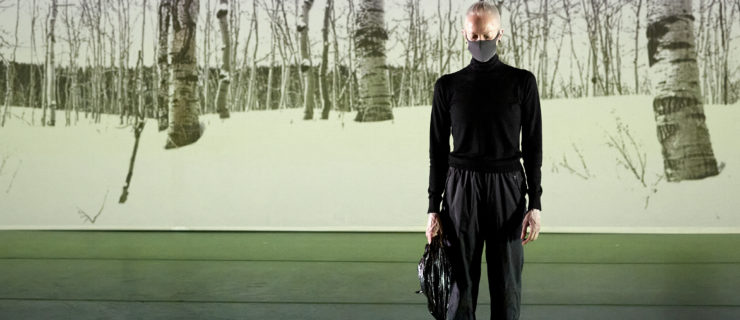 Karole Armitage is a white woman with fair hair slicked back. She wears a long-sleeved black top and black pants and stands in front of an image of a birch forest with snow.