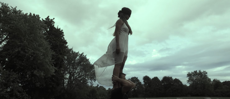 A woman in a sheer white dress stands blindfolded on the shoulders of a person, in a field