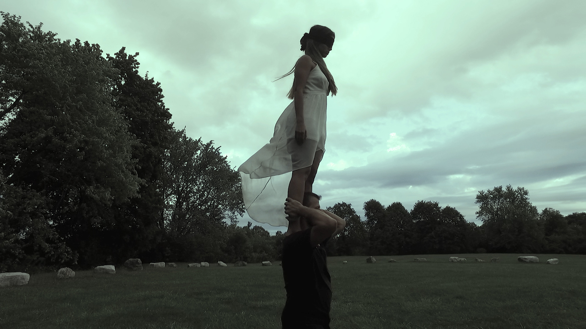 A woman in a sheer white dress stands blindfolded on the shoulders of a person, in a field