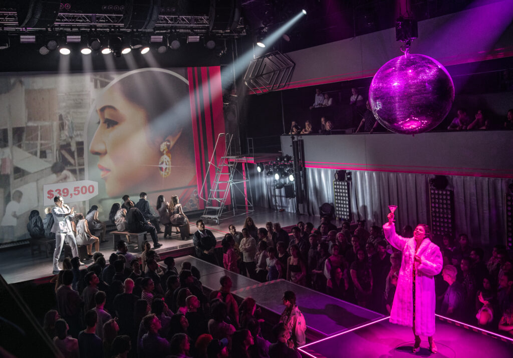 a woman wearing a fur coat holding a drink at the end of the catwalk. a group of performers together update with the audience surrounding them