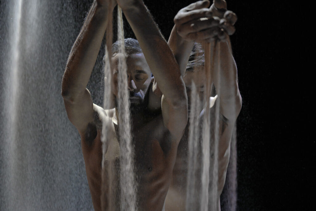 Aakash Odedra and Hu Shenyuan stand close together, one ahead of the other. Their eyes are closed as they raise their hands overhead, cupped fingers allowing streams of sand to fall in front of their faces.